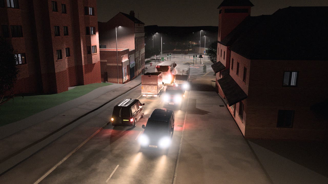 Evening traffic on a street lined with brick buildings, rendered by VeriStreet: vans and lorries with headlights and tail lamps, street lights and signals at a junction ahead.
